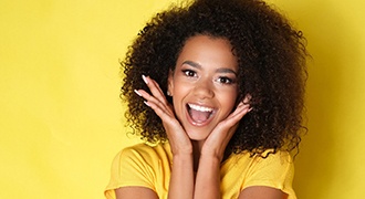 A young woman wearing a yellow blouse holds her face between her hands and smiles